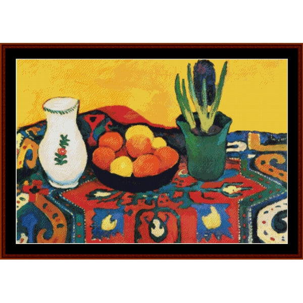 Counted Cross Stitch Pattern – Still Life with Hyacinths by August Macke - Cross Stitch Collectibles - A framed still life painting featuring a white vase with a green floral motif, a bowl of oranges and lemons, and a green potted plant on a colorful patterned tablecloth against a yellow background.