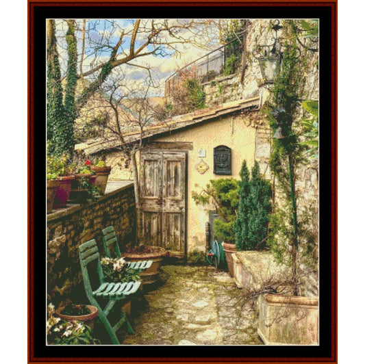 Vintage-style Counted Cross Stitch Pattern by Cross Stitch Collectibles of a quaint outdoor scene with a stone building, green chairs, and potted plants.