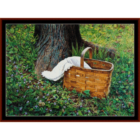 Counted Cross Stitch Pattern: Picnic, Americana - Cross Stitch Collectibles - A framed art print depicting a classic brown woven picnic basket with a white throw blanket, placed on a grassy forest floor next to a large tree trunk.