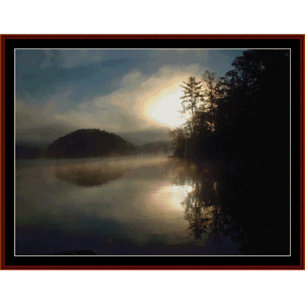 Counted Cross Stitch Pattern – Reflections - Cross Stitch Collectibles - A framed landscape art print depicting a misty lake at sunrise or sunset, with a bright sun glowing over silhouetted trees and a dark hill on the left