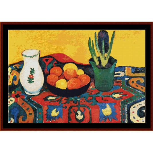 Counted Cross Stitch Pattern – Still Life with Hyacinths by August Macke - Cross Stitch Collectibles - A framed still life painting featuring a white vase with a green floral motif, a bowl of oranges and lemons, and a green potted plant on a colorful patterned tablecloth against a yellow background.